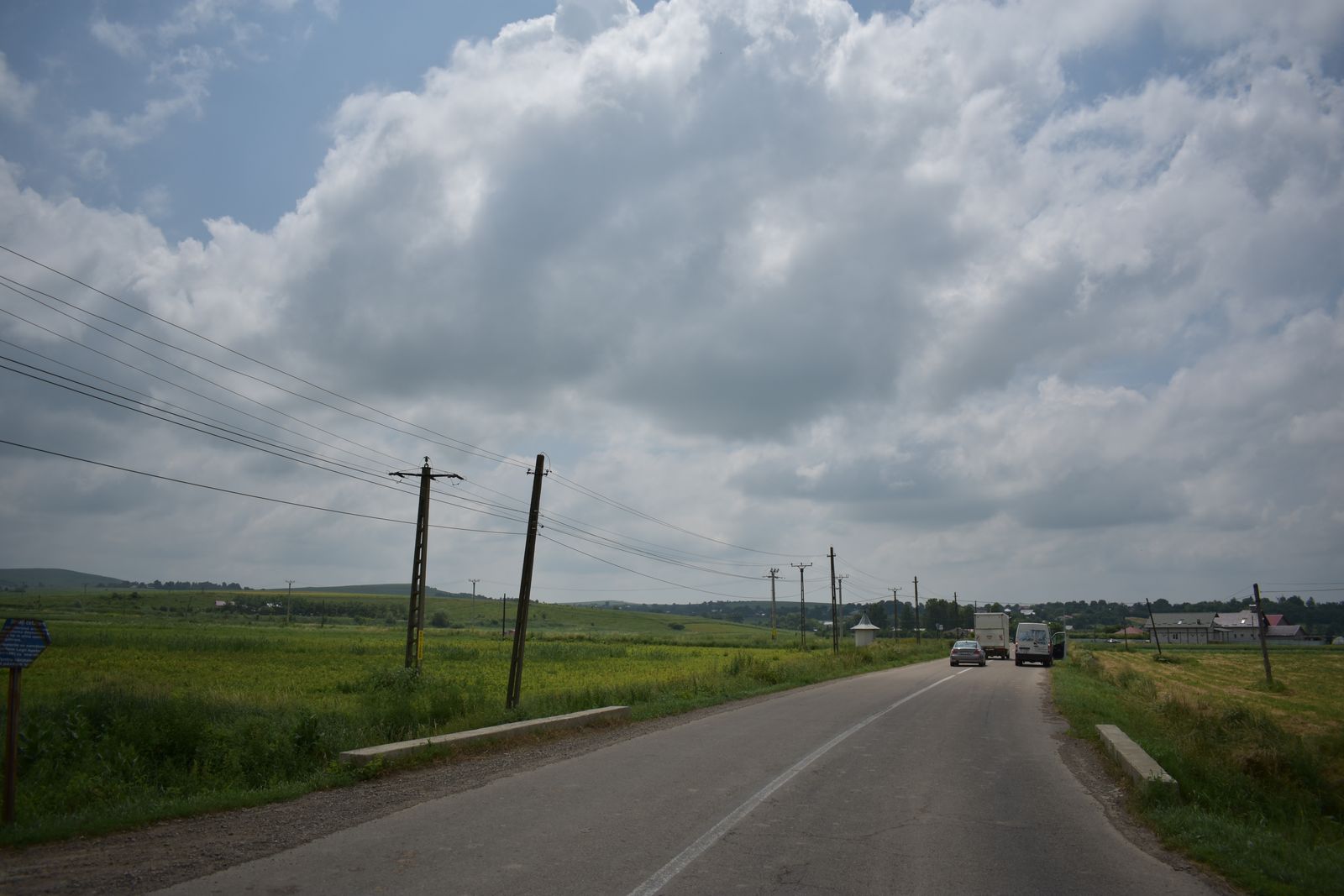 Drum asfaltat între câmpuri, cu stâlpi de rețea electrică și un orizont de dealuri
